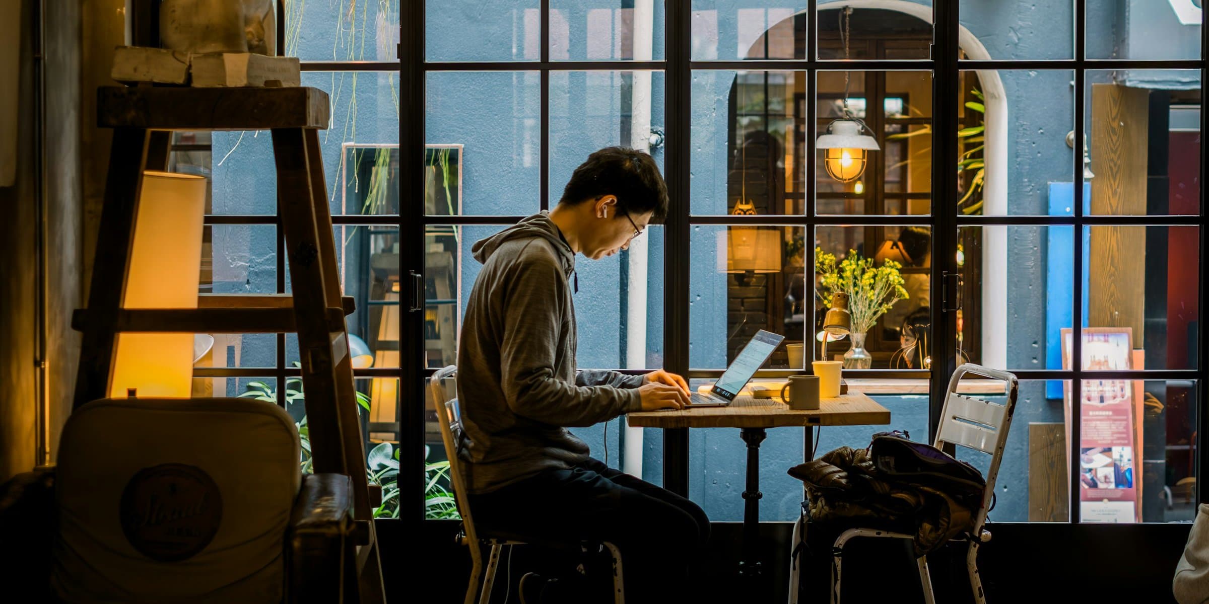 A person working on a laptop at a café table by a window, warm lamplight around them.